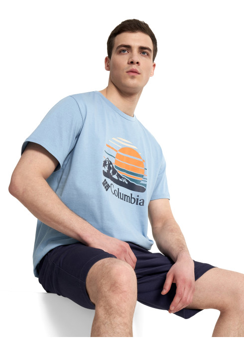Чоловік у блакитній футболці Columbia Path Lake Graphic Tee II сидить на білому кубі на білому тлі